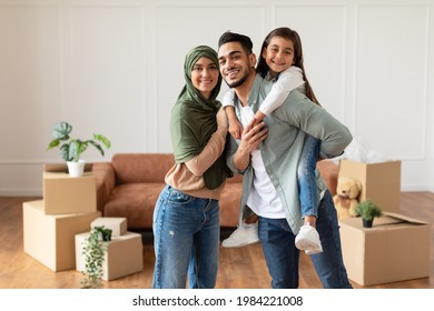 Portrait of excited muslim man, woman in headscarf and girl posing in new apartment, standing in empty living room with cardboard boxes. Cheerful father carrying his daughter on back