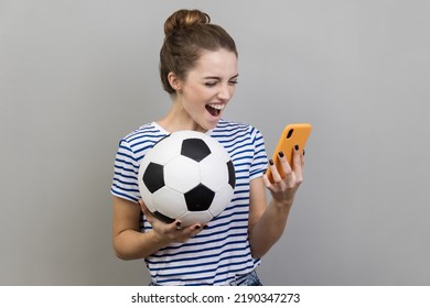 Portrait of excited positive happy woman wearing striped T-shirt holding soccer ball and smart phone, betting and winning, football match. Indoor studio shot isolated on gray background. - Powered by Shutterstock