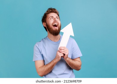 Portrait of excited positive optimistic bearded man holding arrow pointing up looking at camera with smile, growth and increase concept. Indoor studio shot isolated on blue background. - Shutterstock ID 2123698709