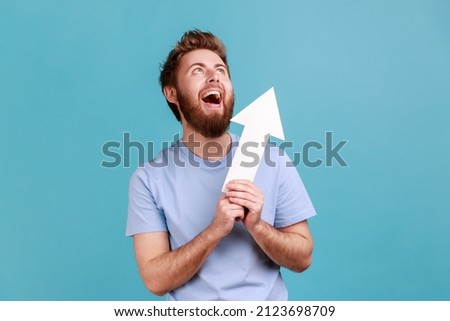 Portrait of excited positive optimistic bearded man holding arrow pointing up looking at camera with smile, growth and increase concept. Indoor studio shot isolated on blue background.