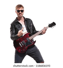 portrait of excited rockstar wearing sunglasses and leather jacket making rock on sign during concert while standing on white background