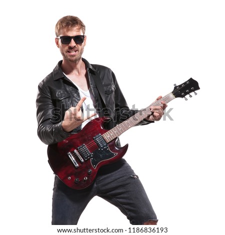 portrait of excited rockstar wearing sunglasses and leather jacket making rock on sign during concert while standing on white background