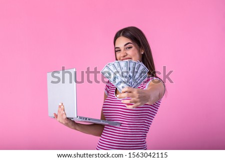 Portrait of an excited satisfied girl holding money banknotes with laptop computer isolated over pink background. Portrait of a cheerful young woman holding money banknotes and laptopcomputer in hands