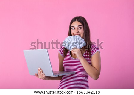 Portrait of an excited satisfied girl holding money banknotes with laptop computer isolated over pink background. Portrait of a cheerful young woman holding money banknotes and laptopcomputer in hands