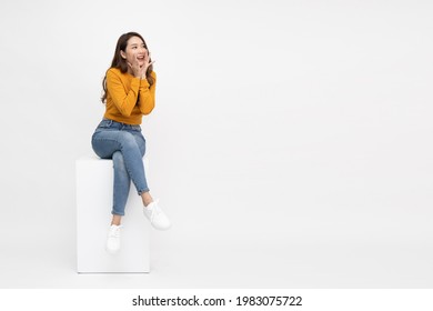 Portrait of excited screaming young asian woman sitting on white box isolated over white background, Wow and surprised concept