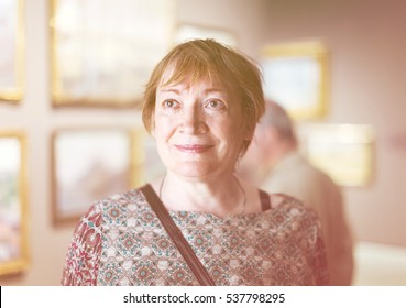 Portrait Of Excited Senior Woman Attentively Looking At Paintings In Art Museum 
