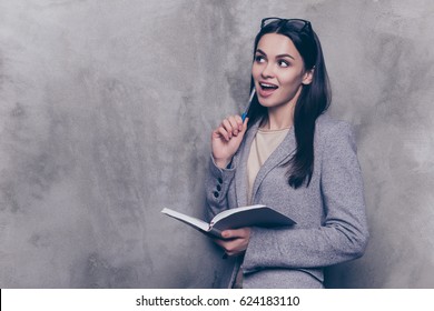 Portrait of excited  smart young successful businesswoman in suit note new idea and do list while standing near gray wall