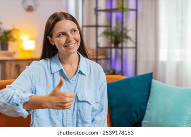 Portrait of excited smiling Caucasian woman looking approvingly at camera showing double thumbs up, like sign, good news, positive feedback. Happy young girl sitting on couch in living room at home. - Powered by Shutterstock