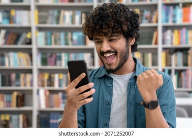 Portrait Of Excited Smiling Indian Student Screaming Holding Mobile Phone And Cheering Standing In Library. Hindu Teen Guy Celebrating Success In Passing Exam Celebrating Victory Or Achievement