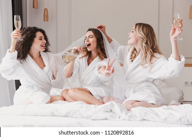 Portrait of excited three women 20s celebrating bachelorette party and drinking champagne in posh apartment or hotel room while bride trying on wedding veil