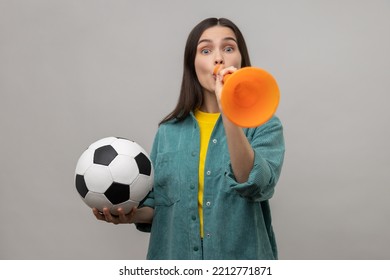 Portrait Of Excited Woman Blowing In Horn Holding Black And White Soccer Ball, Celebrating Victory Of Favourite Football Team, Wearing Casual Jacket. Indoor Studio Shot Isolated On Gray Background.