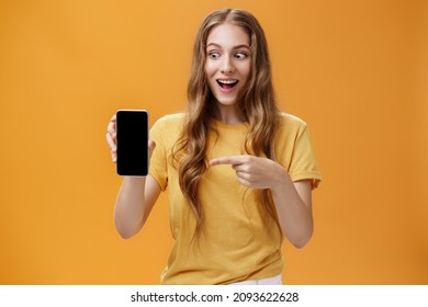 Portrait of excited woman feeling amazed holding awesome new smartphone in hand pointing at cellphone screen popping eyes like crazy at device being charmed with cool technology product