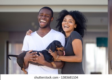 Portrait Of Excited Young African American Couple Posing With Domestic Pet Outside New Family House, Happy Black Husband And Wife With Cute Dog Laugh Glad To Buy First Home Together. Ownership Concept