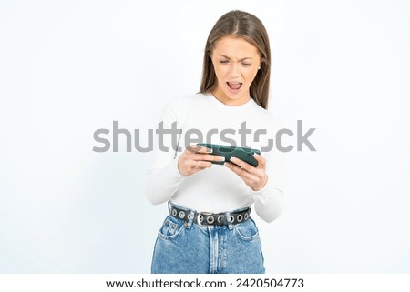 Portrait of an excited Young beautiful woman standing over white studio background playing games on mobile phone.