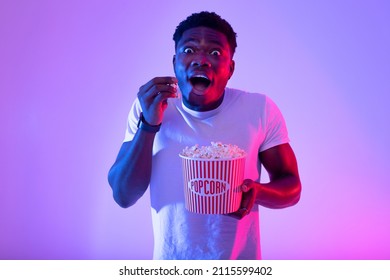 Portrait Of Excited Young Black Guy Eating Popcorn From Bucket, Watching Movie In Cinema Or On TV, Neon Light. Cheerful African American Man Enjoying Cool Film, Having Tasty Snack