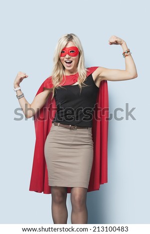 Portrait of excited young blond woman in superhero costume flexing arms over light blue background