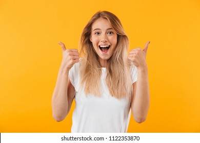 Portrait of an excited young blonde girl showing two thumbs up isolated over yellow background