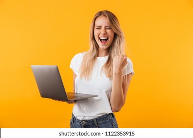 Portrait of an excited young blonde girl holding laptop computer and celebrating success isolated over yellow background