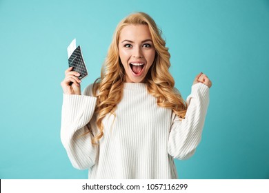 Portrait of an excited young blonde woman in sweater holding passport with flying tickets isolated over blue background