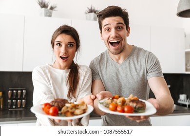 Portrait Of An Excited Young Couple Holding Dinner Plates With Food While Standing At The Kitchen