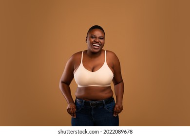 Portrait of excited young curvy African American woman smiling and laughing looking at camera wearing bra and jeans standing isolated over brown wall. Cheerful chubby lady posing, hearing good news