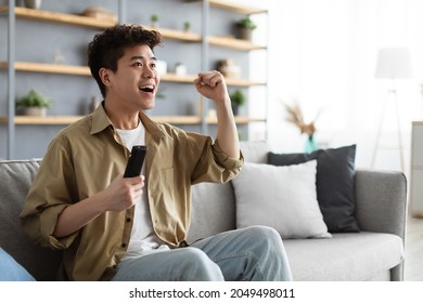 Portrait Of Excited Young Guy Watching Football Match, Shaking Clenched Fist. Happy Asian Man Relaxing Sitting On Couch Cheering Favorite Team Enjoying Game On TV At Home Holding Remote Controller