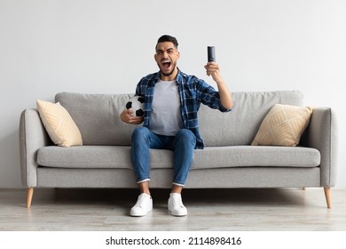 Portrait of excited young guy watching football match, raising clenched fist. Emotional man sitting on couch cheering favorite team enjoying game goal on TV at home holding remote controller and ball