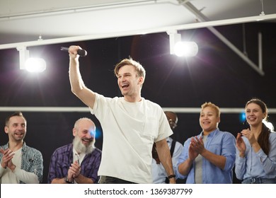 Portrait of excited young man holding microphone standing on stage in spotlight  with people applauding in background, copy space - Powered by Shutterstock