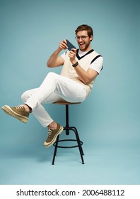Porträt eines aufgeregten jungen Mannes, der auf einem Hocker sitzt und Spiele auf dem Handy spielt, einzeln auf blauem Hintergrund.