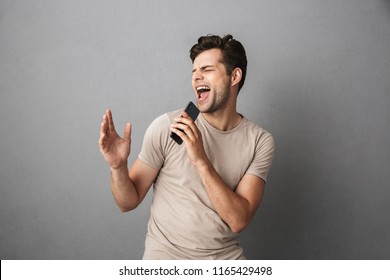 Portrait of an excited young man in t-shirt isolated over gray backgound, holding blank screen mobile phone, singing