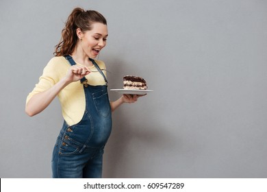 Portrait Of An Excited Young Pregnant Woman Eating Chocolate Cake Isolated On A Gray Background