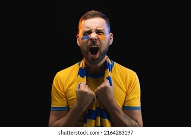Portrait of excited young soccer supporter man in yellow-blue t-shirt and scarf and painted face, cheering for his favourite team. Isolated over black background. - Shutterstock ID 2164283197