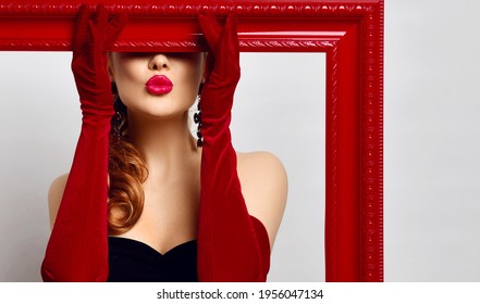 Portrait excited young woman artist in black off  shoulder tight dress  red elbow  length gloves holding big red empty picture frame covering her eyes   sending  blowing kiss