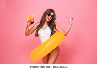 Portrait of an excited young woman dressed in swimsuit posing with inflatable ring and holding a cocktail isolated over pink background