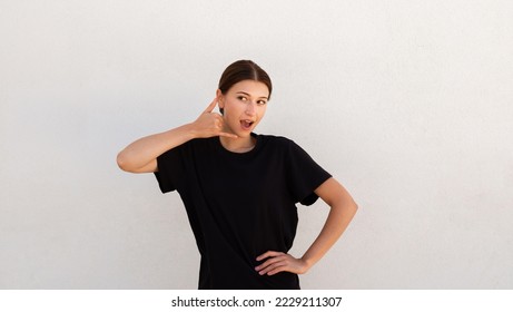 Portrait of excited young woman making mobile phone gesture over white background. Caucasian woman wearing black T-shirt asking to call her back. Mobile communication and feedback concept - Shutterstock ID 2229211307