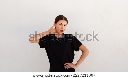 Portrait of excited young woman making mobile phone gesture over white background. Caucasian woman wearing black T-shirt asking to call her back. Mobile communication and feedback concept