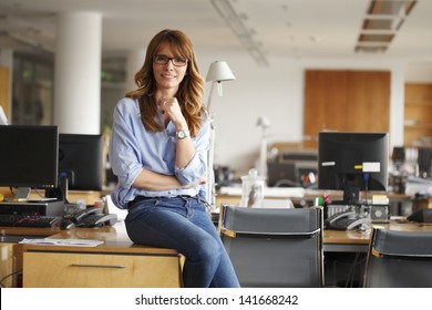 Portrait of an executive professional mature businesswoman sitting on office desk in casual and smiling. Shallow focus.