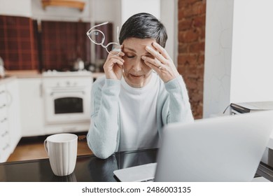 Portrait of exhausted female entrepreneur of tired senior businesswoman in warm sweater rubbing eye after intense work on laptop, working from home remotely, holding glasses in hands - Powered by Shutterstock