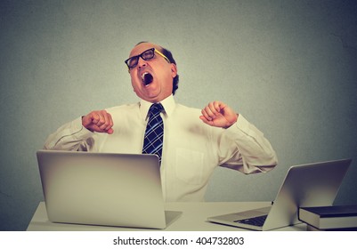 Portrait Of An Exhausted Middle Aged Business Man Yawning At Work In Office Sitting At His Desk Table With Many Laptop Computers. Lack Of Sleep Productivity Concept. Sleep Deprivation Long Work Hours