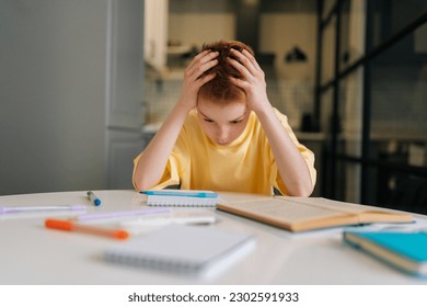 Portrait of exhausted pupil boy tired from studying holding head head with hands sitting at desk with paper copybook, looking down. Frustrated child schoolboy doing homework at home. - Shutterstock ID 2302591933