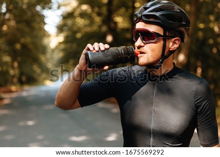 Portrait of exhausted sportsman in black helmet and mirrored glasses drinking cold water from sport bottle while standing in green forest. Mature man having break during long ride.