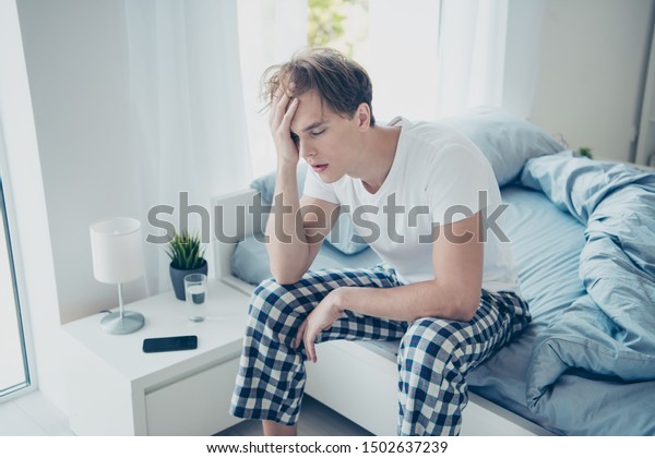 Portrait of exhausted unhappy man after\
hangover party have terrible fatigue migraine catch cold suffer sit\
on bed wear checkered plaid pajama in house\
indoors
