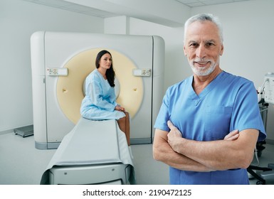 Portrait Of Experienced Doctor Standing In Medical Radiology Room With CT Scanner, Female Patient Sitting Behind His Waiting For CT Scan