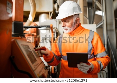 Portrait of an experienced industrial engineer or senior factory worker operating automated machine in production hall. Factory interior in background.