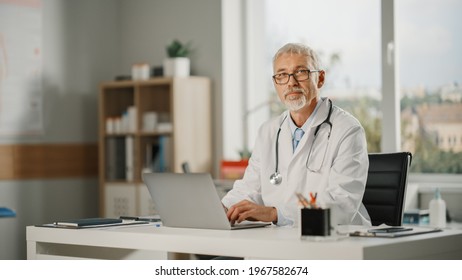 Portrait Of An Experienced Middle Aged Male Doctor Wearing White Coat Working On Laptop Computer At His Office. Senior Medical Health Care Professional Looks At The Camera And Gently Smiles.