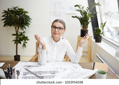 Portrait Of Experienced Middle Aged Senior Female Architect In Stylish Glasses And White Blouse Sitting At Her Workplace With Drawings Of Architectural Project On Desk, Rejoicing At Well Done Work