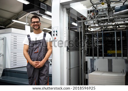 Portrait of an experienced typographer standing by modern printing machine in print house.
