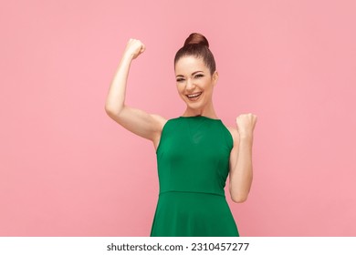 Portrait of extremely happy positive woman standing with raised clenched fists, winning lottery, celebrating her success, wearing green dress. Indoor studio shot isolated on pink background. - Shutterstock ID 2310457277
