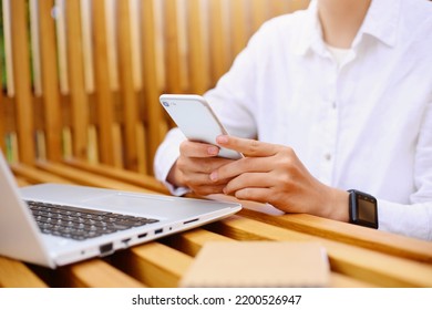 Portrait of faceless woman wearing white shirt sitting at wooden table near laptop and using cell phone, browsing internet, checking social network or answering message.