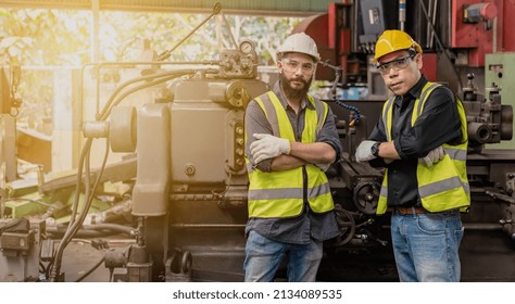 Portrait Of Factory Engineer And Asian Co-worker In Safety Uniform With Protective Hard Helmet And Vest Standing In Industrial Manufacturer Factory With Heavy Machinery Background, Technicians Workers